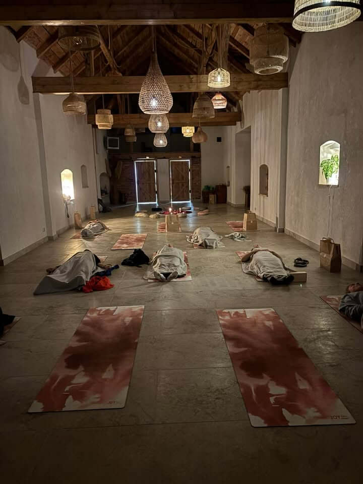 Sala warsztatowa wieczorem podczas joga nidry – uczestniczki leżą na matach przykryte kocami, drewniany strop oświetlony plecionymi lampami w stylu boho