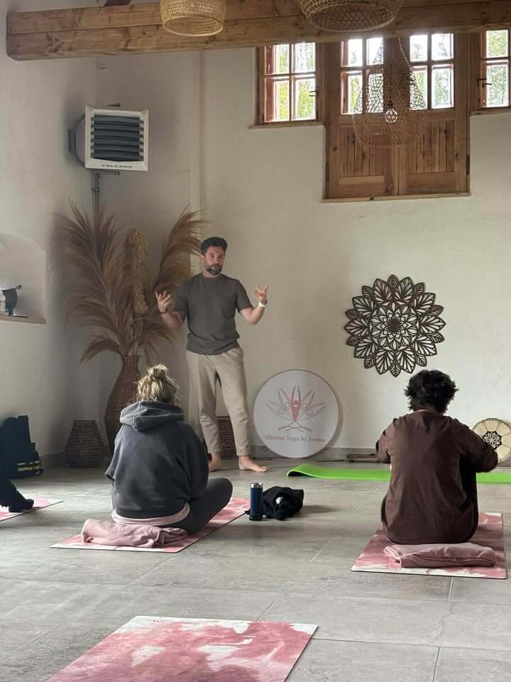 Prowadzący mężczyzna w szarej koszulce wyjaśnia ćwiczenie dwóm uczestniczkom siedzącym na różowych matach; w tle drewniana mandala i logo Ahimsa Yoga by Iwona